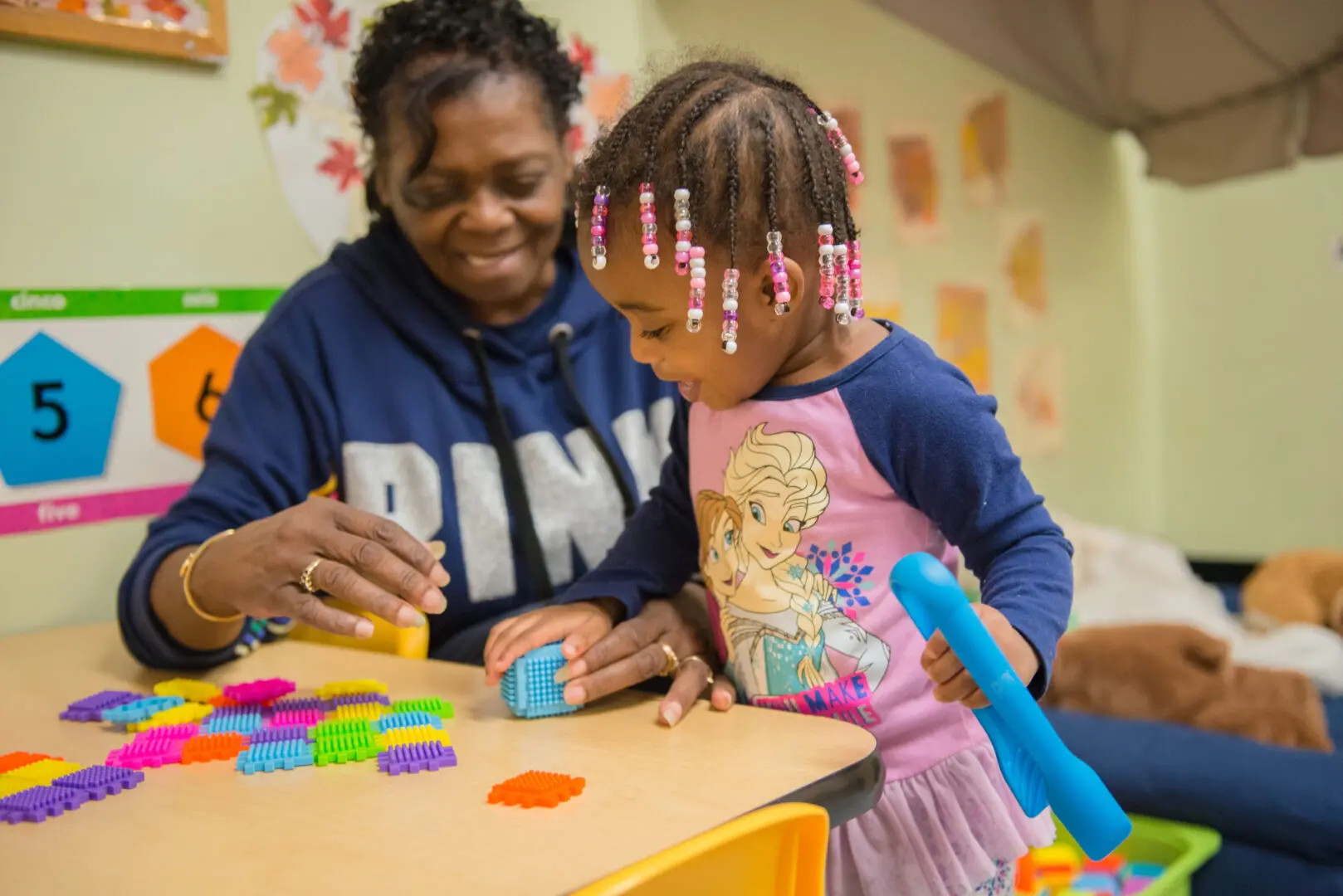 Child and adult playing with colorful toy blocks at a table. The child is holding a blue item, and the adult is smiling and assisting. Educational posters are on the wall in the background.