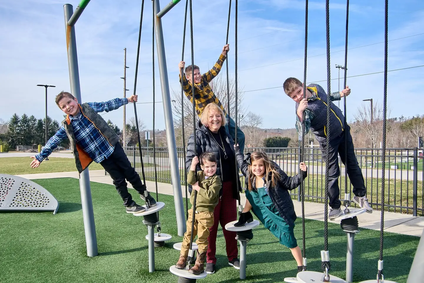 Children and an adult are playing on a modern playground structure with circular platforms and ropes, set against a backdrop of trees and a clear sky.