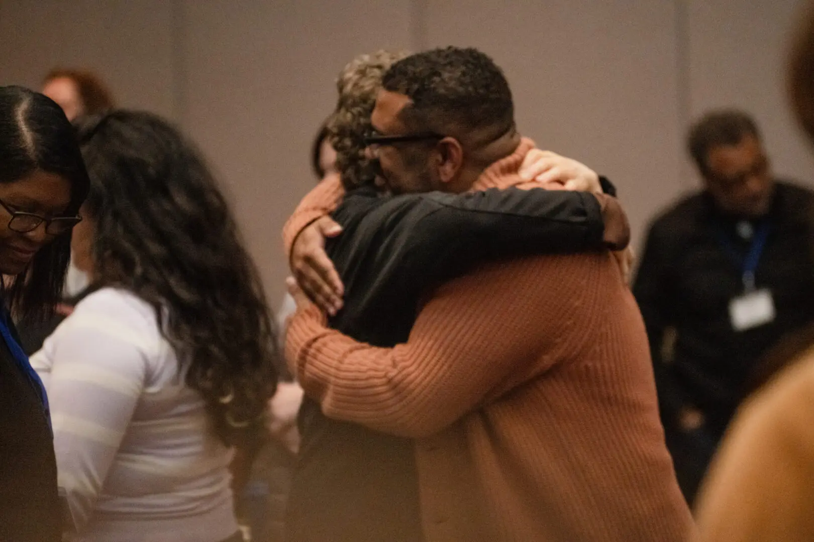 Two people hugging warmly in a room with several others. Some individuals wear name tags and lanyards.