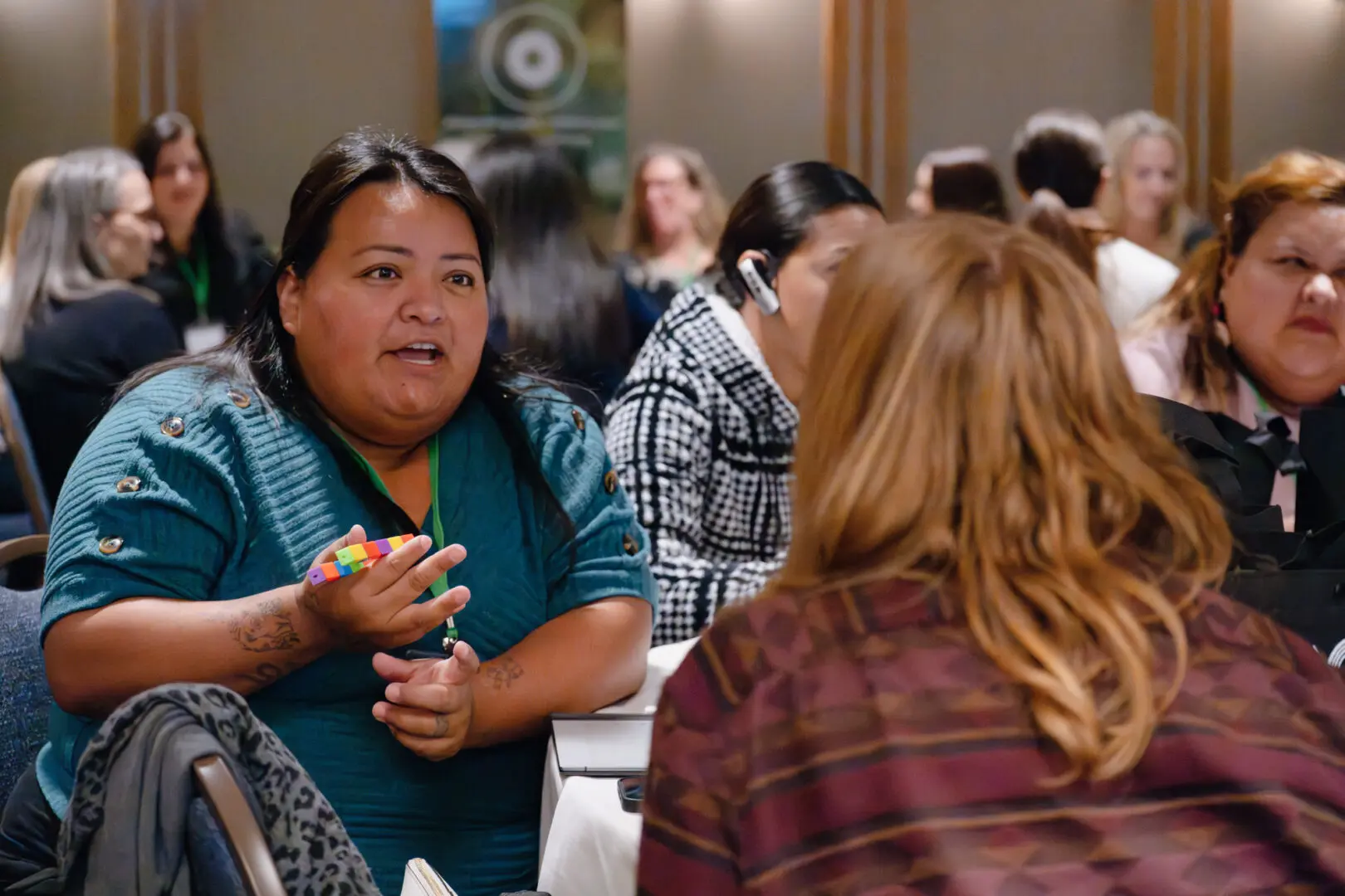 A woman in a teal shirt holds a multicolored cube, engaged in conversation with another woman. Several people are seated around them at tables in a meeting room.
