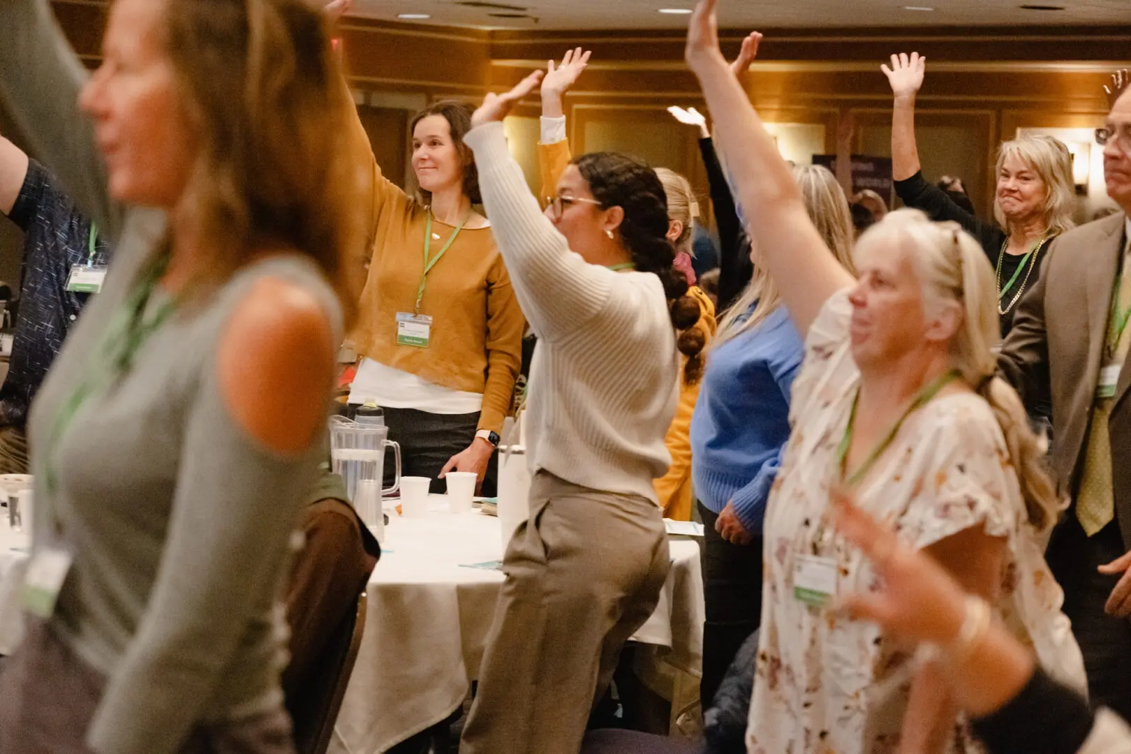A group of people stand and raise their hands in a conference room, appearing engaged in an activity. They wear name tags and casual attire.