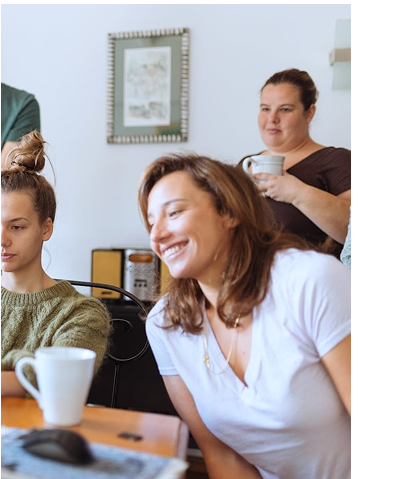 Three women are gathered around a table with coffee mugs, looking at a computer screen and smiling in a casual indoor setting.