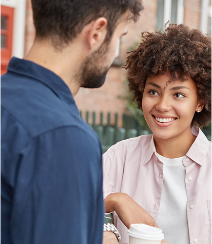 Two people stand outside having a conversation. The woman is smiling and holding a coffee cup, while the man faces her with his back to the camera.