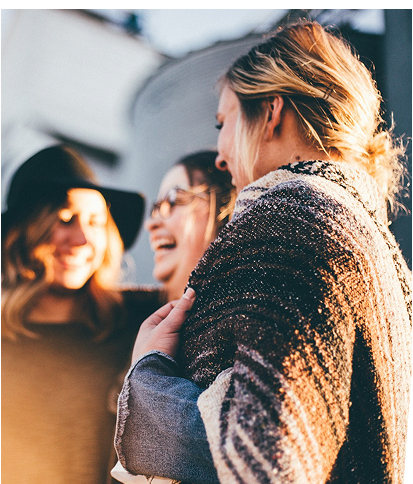 Three women stand close together outdoors, smiling and talking, with one wrapped in a blanket.