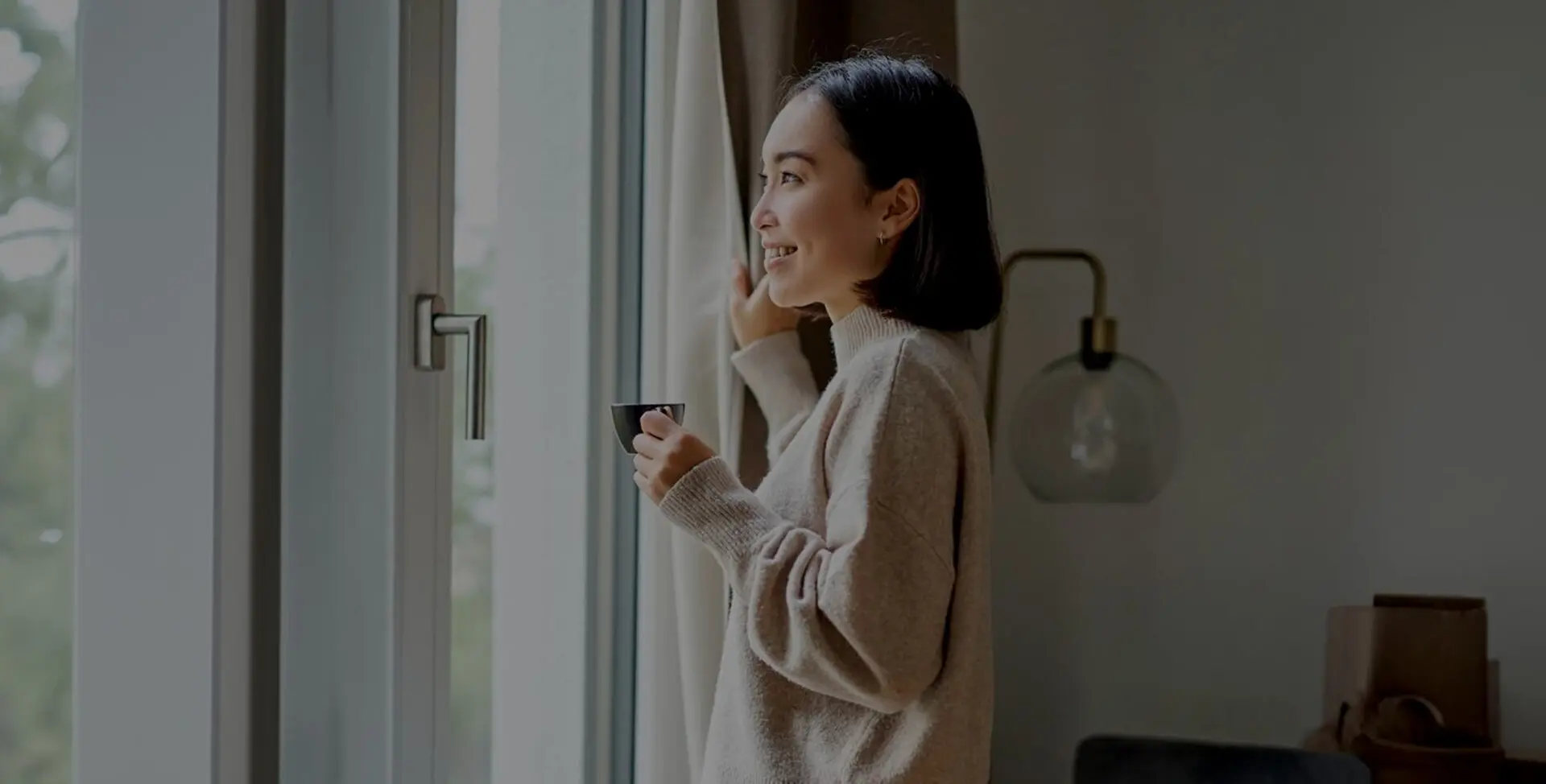 A woman stands by a window holding a cup, looking outside with a slight smile in a softly lit room.