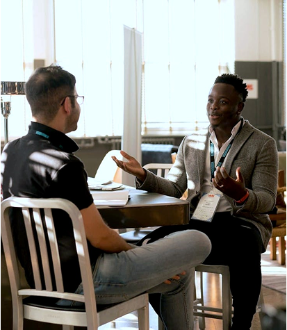 Two men sit across from each other at a table in a bright room, engaged in conversation. One gestures with his hands while the other listens attentively.