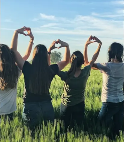 Four people stand in a green field with their backs to the camera, raising their arms to make heart shapes with their hands against a blue sky.