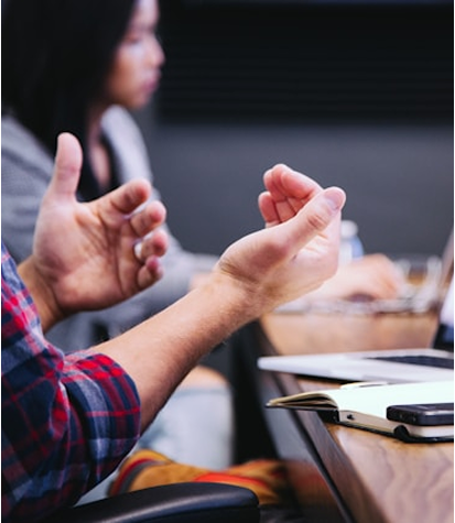 A close-up of a person gesturing with their hands during a meeting, with another person blurred in the background and a notebook and laptop on the table.