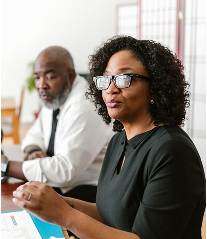 Two professionals are seated at a table in a meeting room; the woman in the foreground is speaking, while the man in the background is listening attentively.