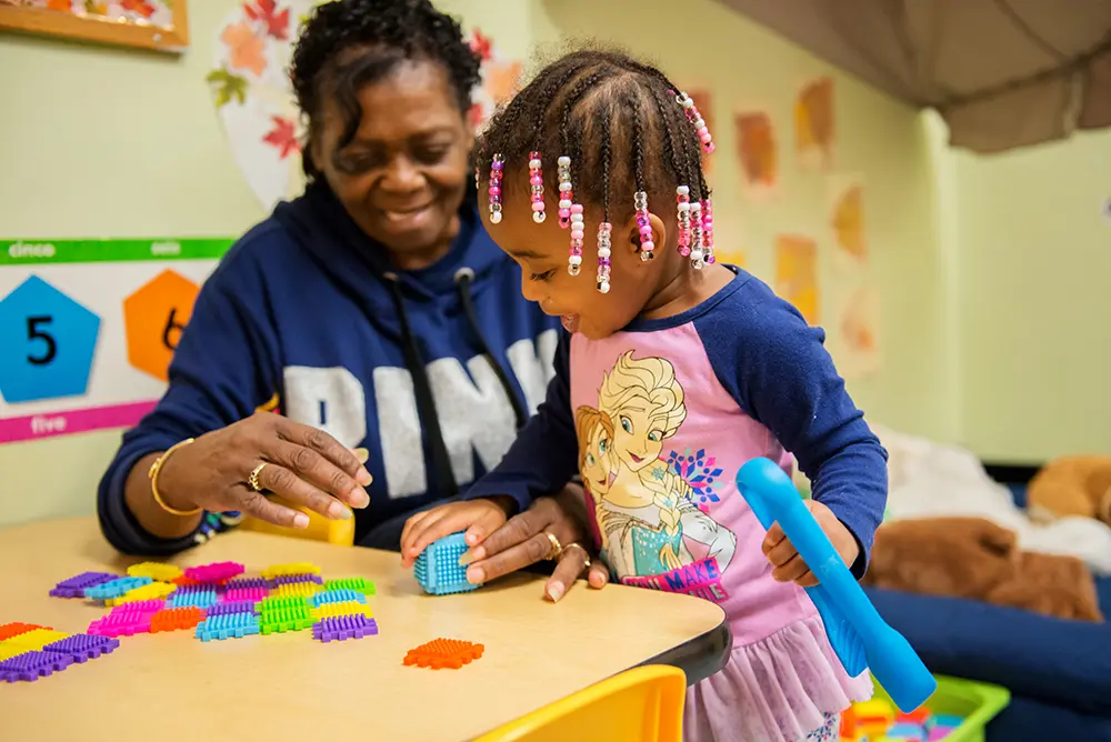 An adult and a child play with colorful interlocking blocks at a table in a classroom decorated with fall-themed artwork.