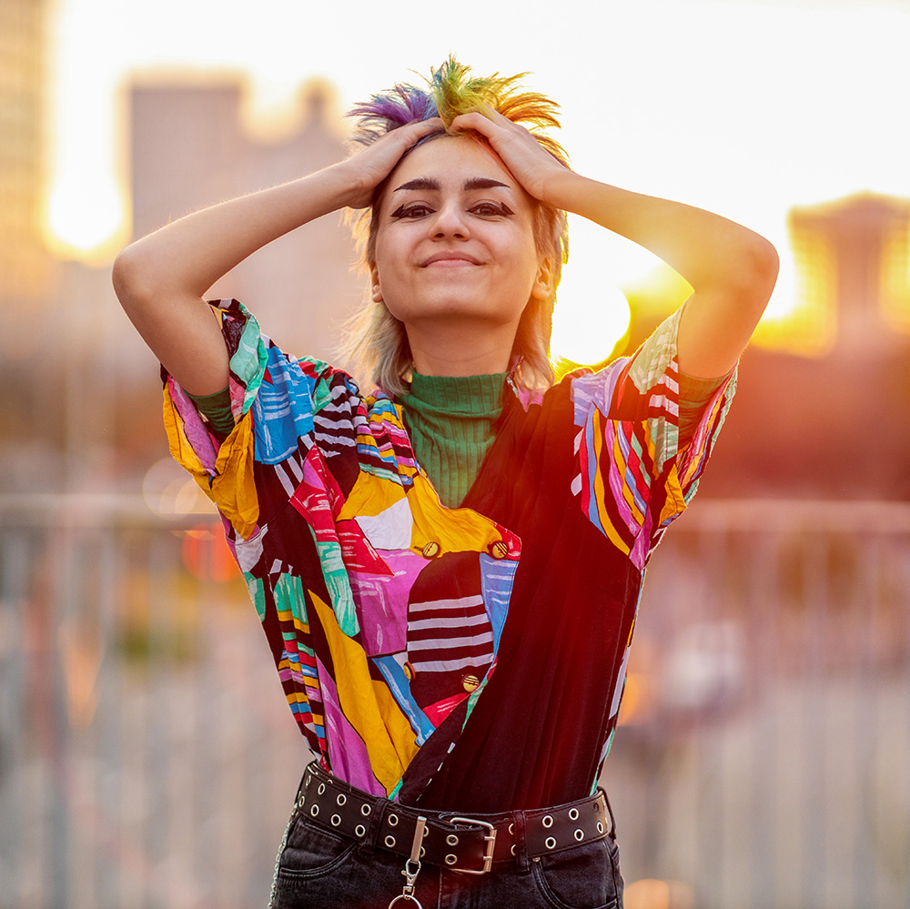 A woman wearing a colorful shirt is posing with her hands on her head.