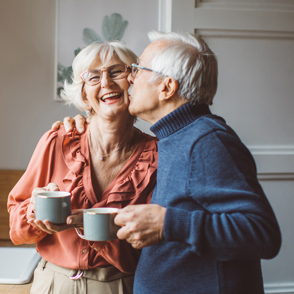 An older couple kissing while holding coffee mugs.