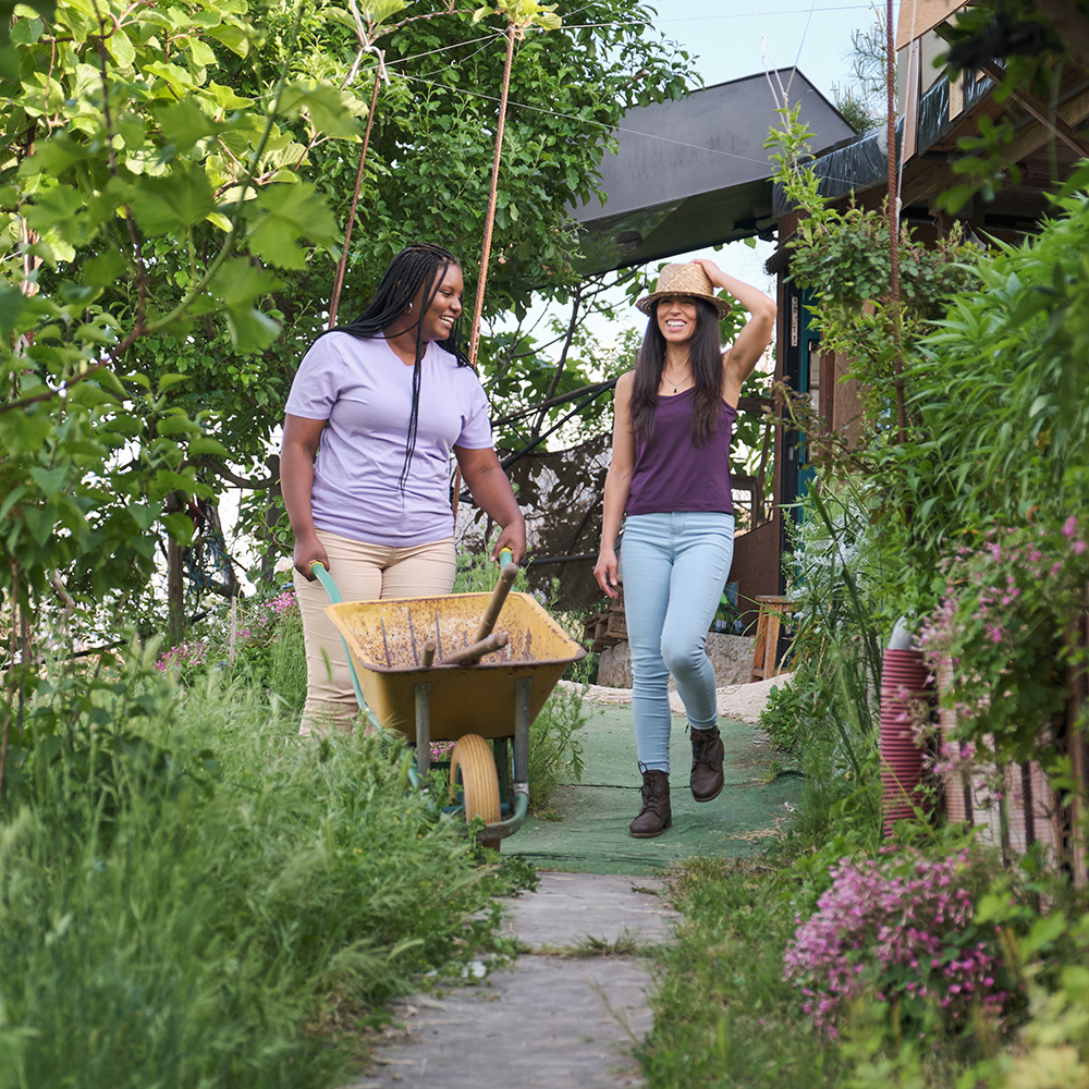 Two women pushing a wheelbarrow through a garden.