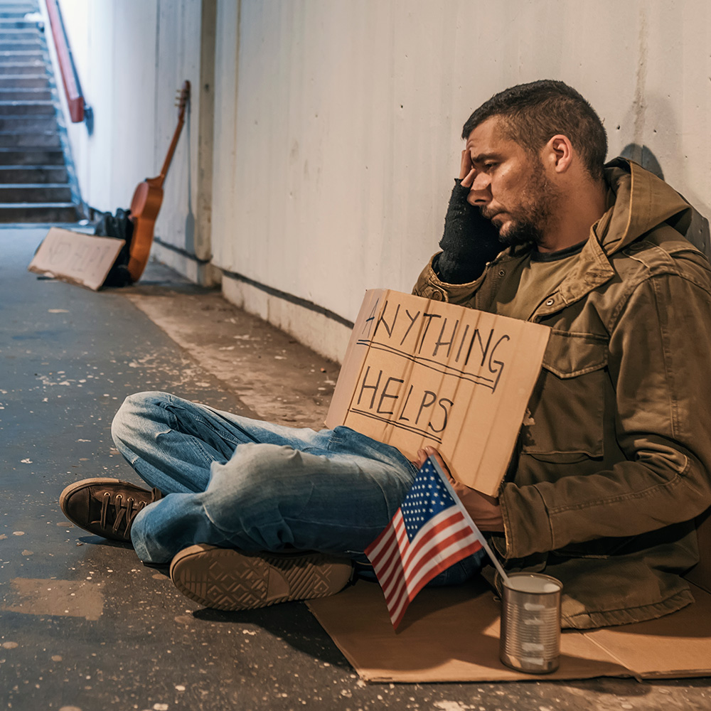 A homeless man with an american flag holding a sign.