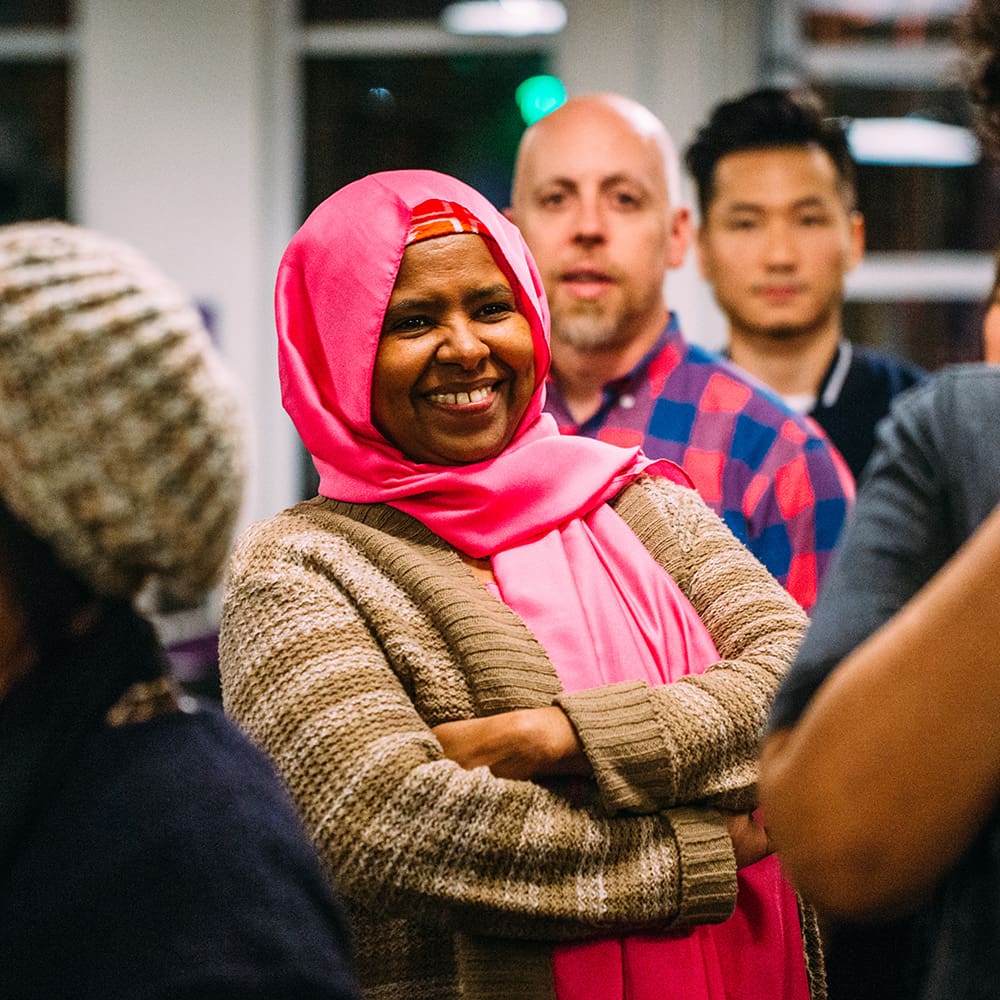 Woman in a pink hijab smiling with crossed arms, standing in a group of people in a casual indoor setting.