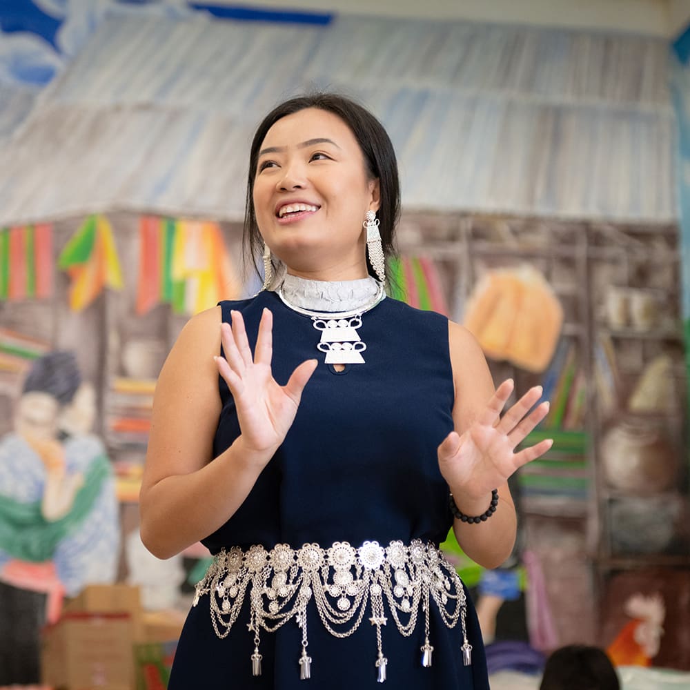 A woman in traditional hmong attire speaking animatedly in a classroom decorated with children's artwork.