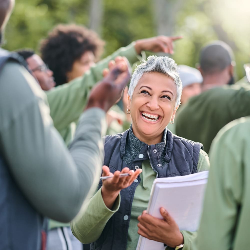 A smiling person with short gray hair holds a stack of papers while engaging with a group of people in an outdoor setting. Several individuals in the background are pointing and gesturing.