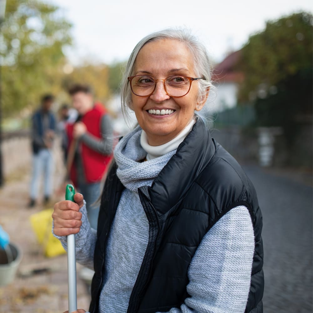 A smiling elderly woman with gray hair and glasses holds a broom outdoors. Other people are visible in the blurred background.