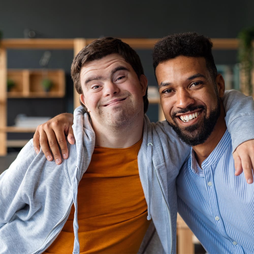 Two men smiling, with one having his arm around the other. They are indoors, dressed casually, and appear to be enjoying each other's company. Shelves and plants are visible in the background.