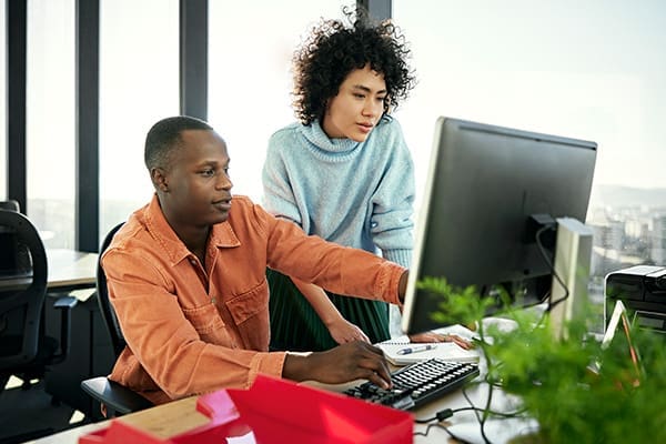 Two people working together at a desk, looking at a computer screen. The person seated is using the keyboard, while the person standing observes. They are in a well-lit office environment.