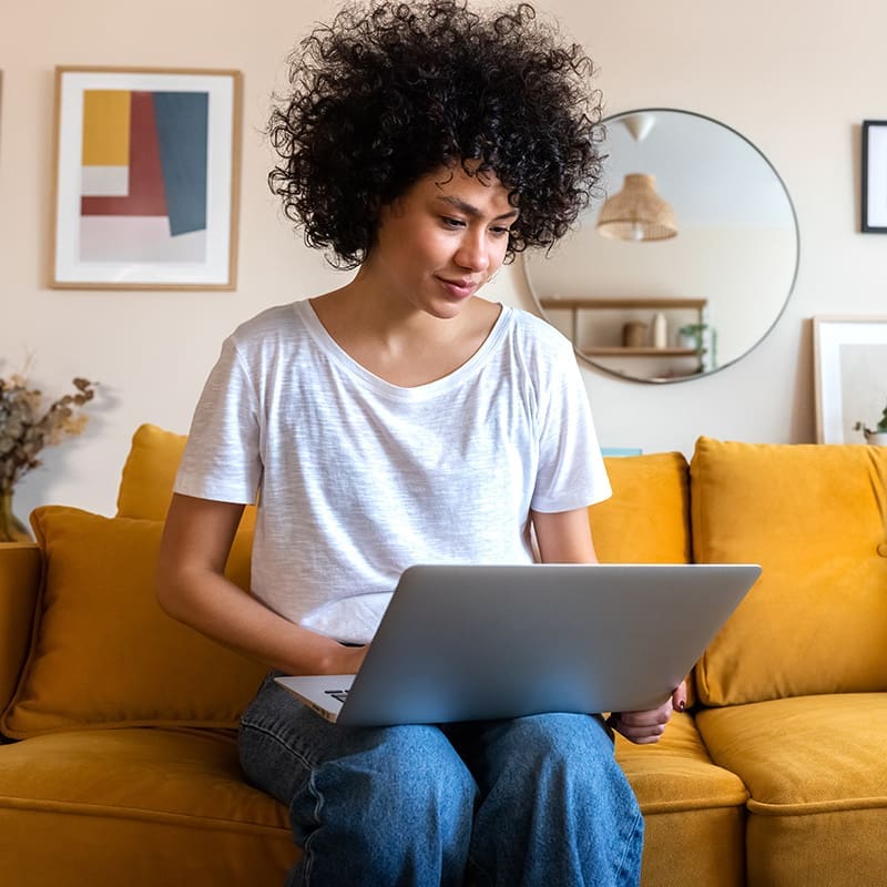 A person with curly hair sits on a yellow couch, using a silver laptop. The room has modern decor, including framed artwork, a round mirror, and various home accessories.