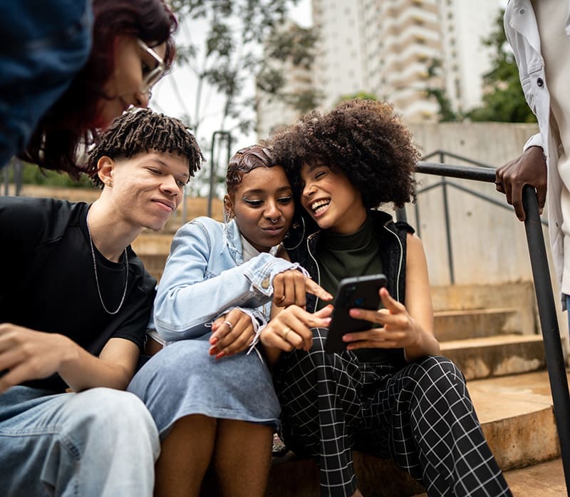 Three people sit on outdoor steps, smiling and looking at a smartphone one of them is holding. Trees and buildings are visible in the background.