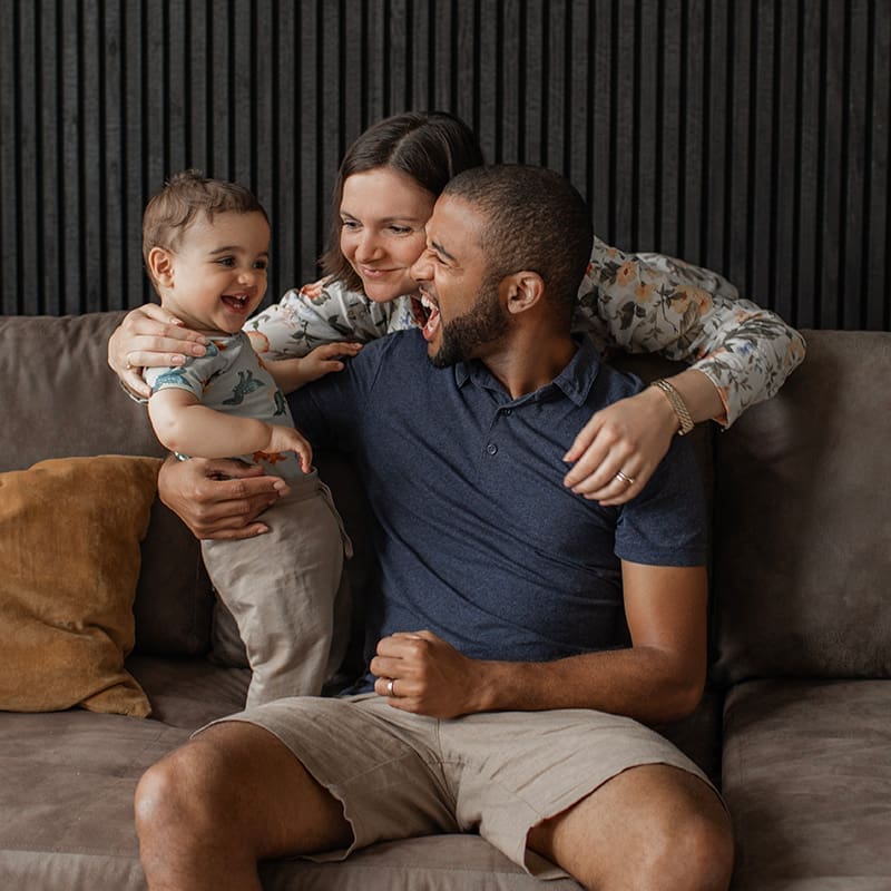 A family of three sits on a couch smiling and interacting. The mother hugs the father and child, who are both laughing.