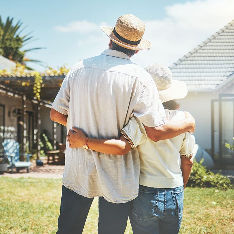 A couple wearing hats, standing arm-in-arm, looks at a house with a garden on a sunny day.