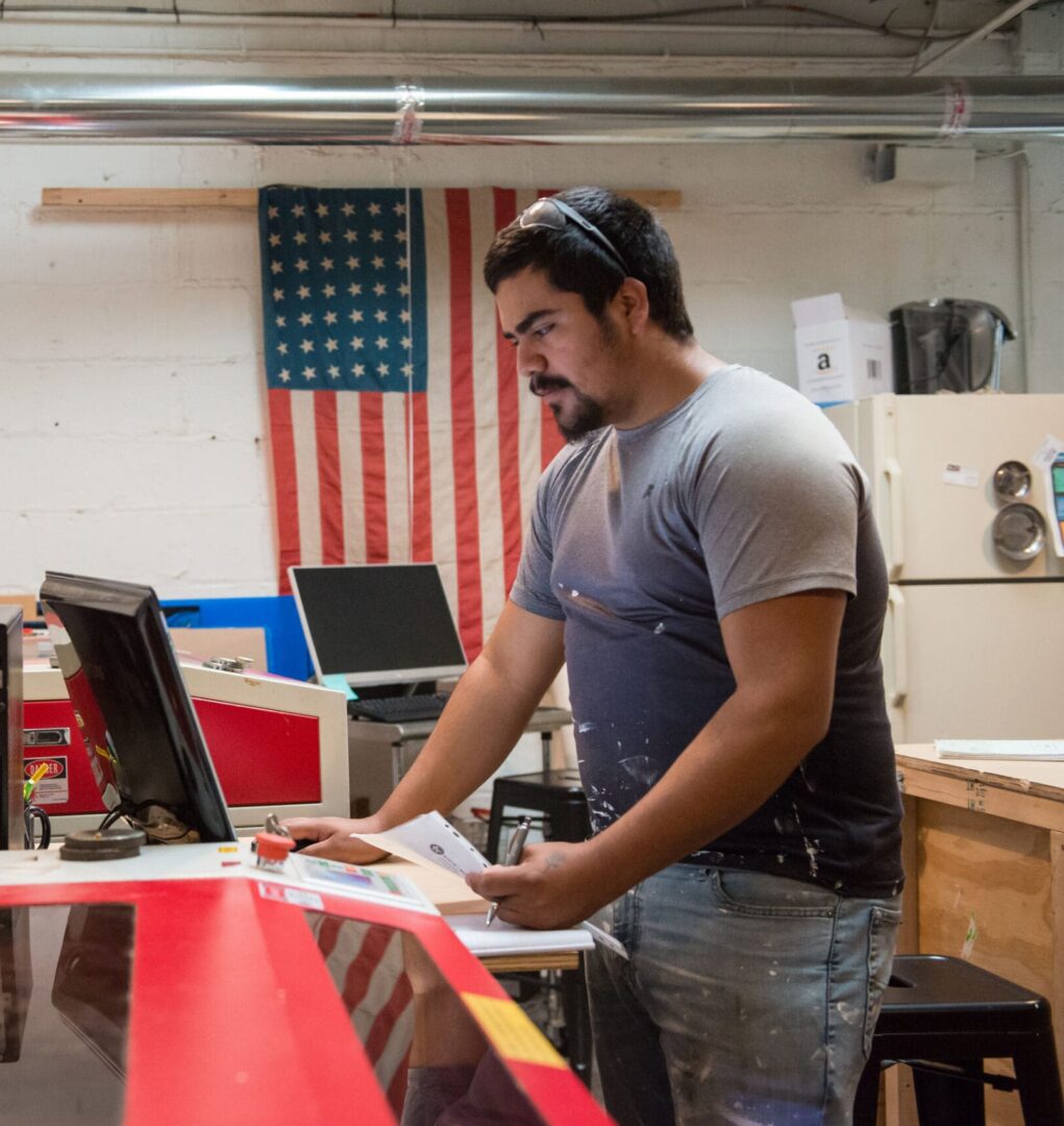 A man in a workshop operates a large machine while looking at a computer screen. An American flag hangs in the background.